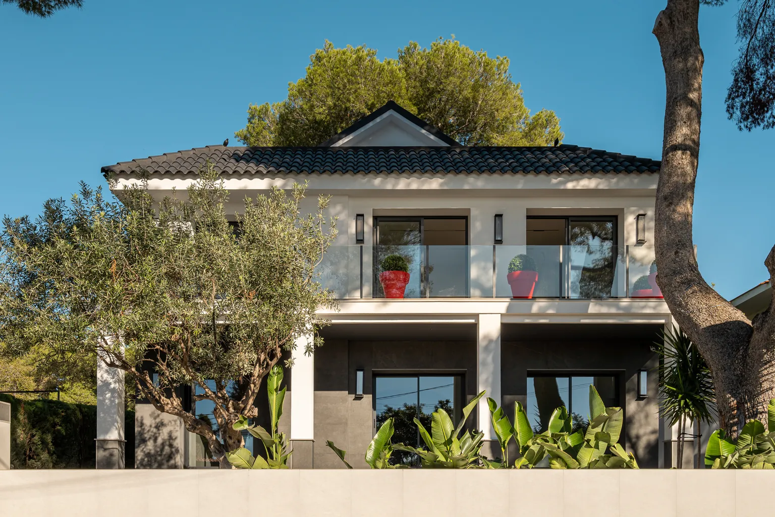 Renovated façade of a detached house among pine trees in Castelldefels, with a black tiled roof, glass balustrades and a dark-clad ground floor
