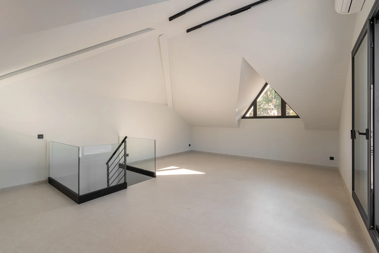 Attic space with a plasterboard ceiling, magnetic lighting tracks, a triangular window and the glass balustrade of the staircase