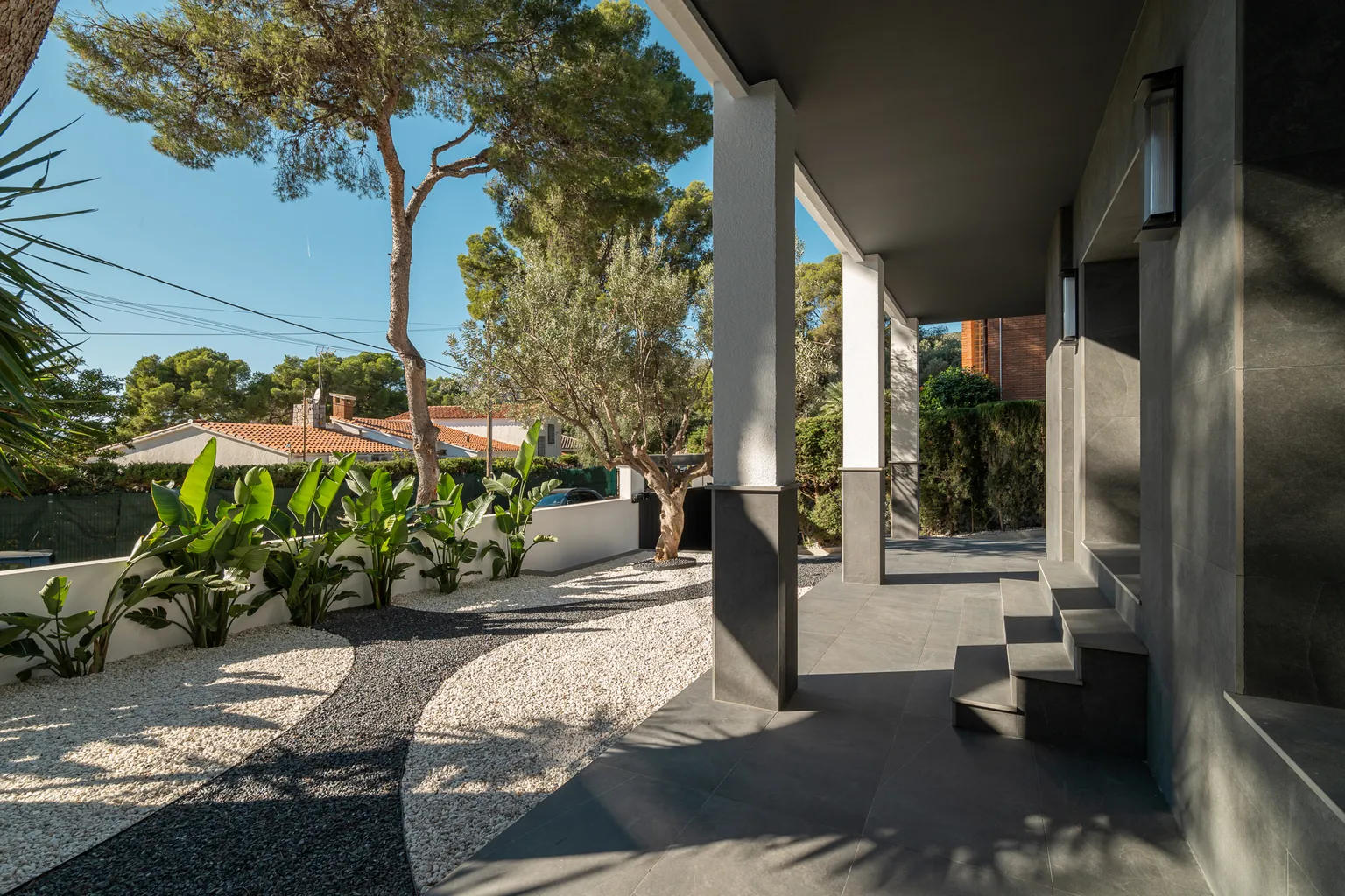 Porch of the house with white columns, dark large-format cladding and a gravel garden with an olive tree and banana plants