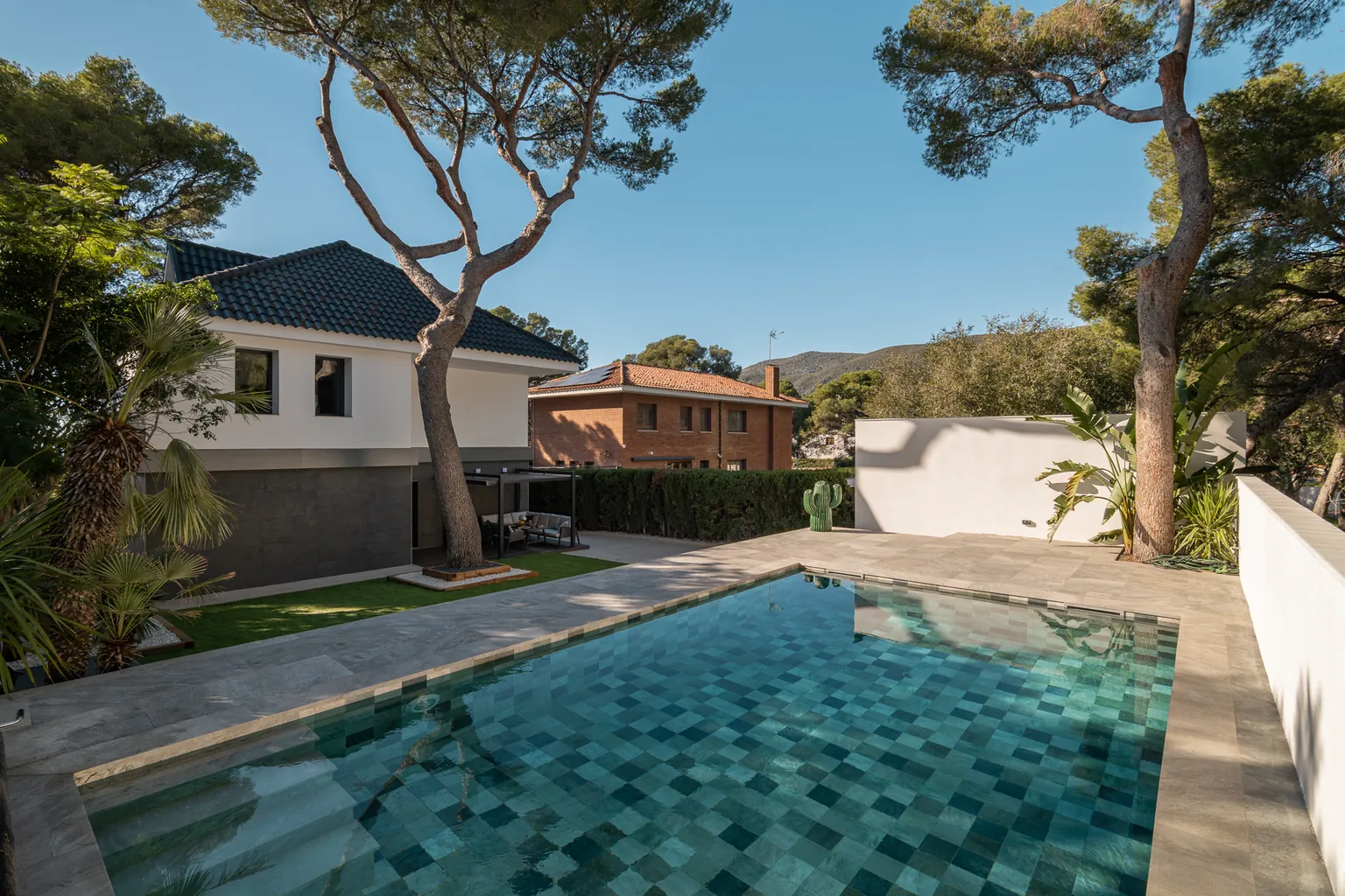 New swimming pool tiled in shades of green and blue, stone-effect outdoor paving and the house with its black roof among pine trees