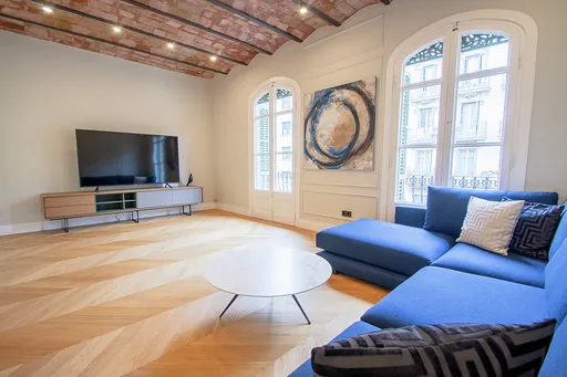 Living room with an L-shaped blue sofa, a white coffee table, a TV unit, an abstract painting and a brick-vault ceiling with spotlights