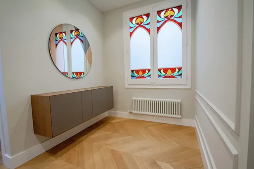 Restored wooden windows with colourful floral stained glass, a round mirror, a floating sideboard and a white radiator on herringbone parquet