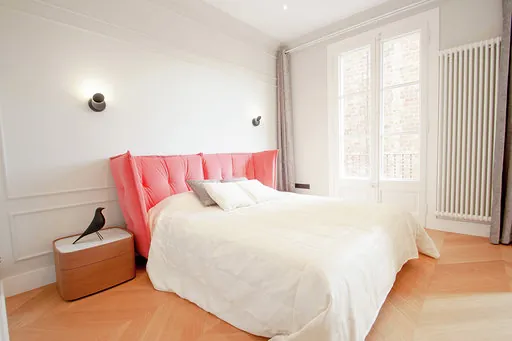 Bedroom with a coral pink upholstered headboard, wall mouldings, black wall lights, a wooden bedside table and a balcony