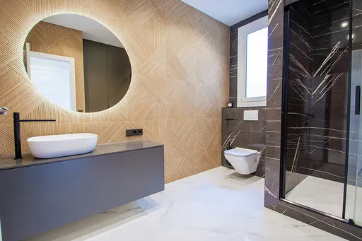 Bathroom with wood-look tiles in a geometric pattern, a round backlit mirror, a basin on a grey top, black tapware and a shower in black marble-effect porcelain