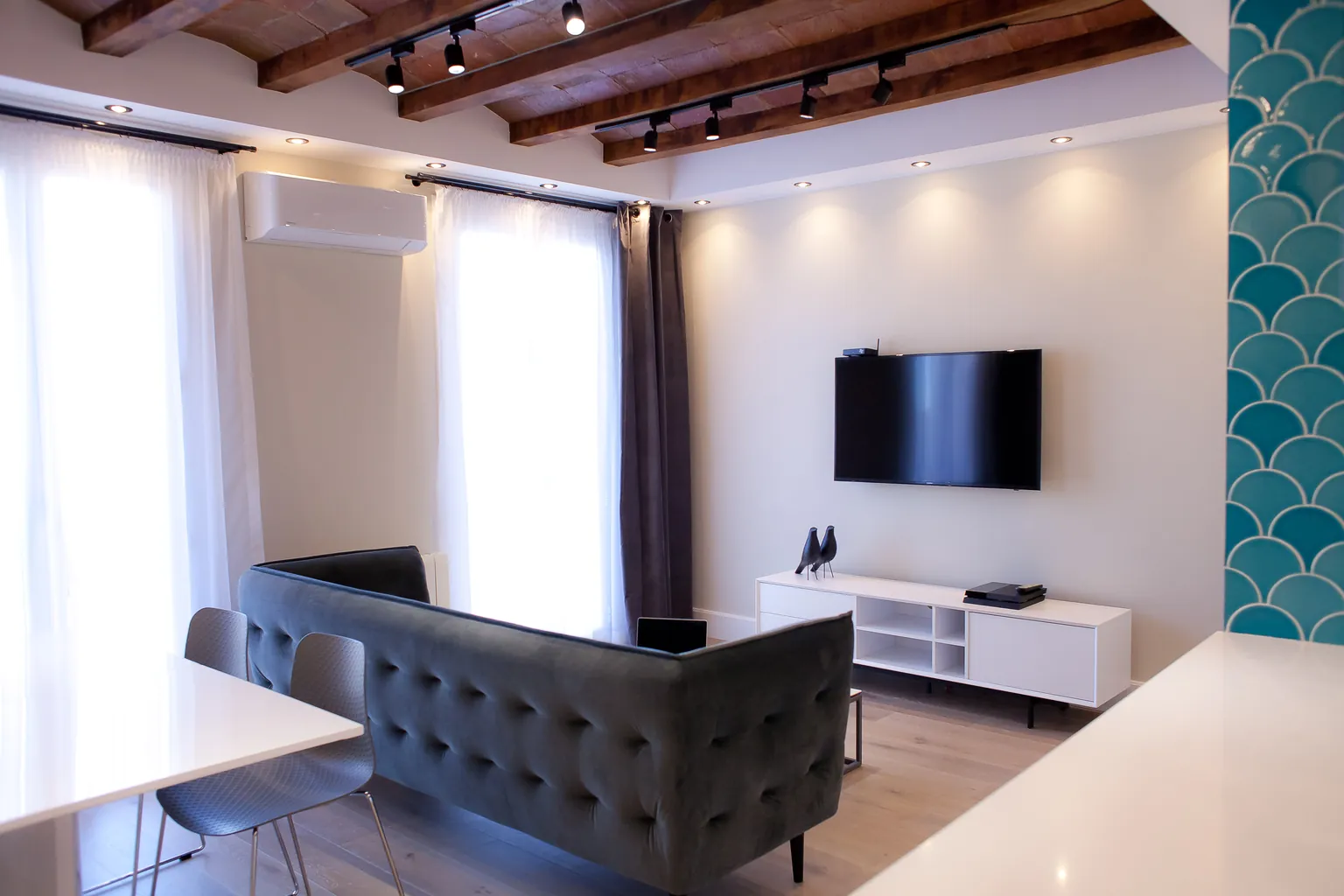Salón con sofá gris capitoné, televisor, mueble blanco, cortinas y techo de vigas de madera con bóvedas de ladrillo e iluminación en carriles