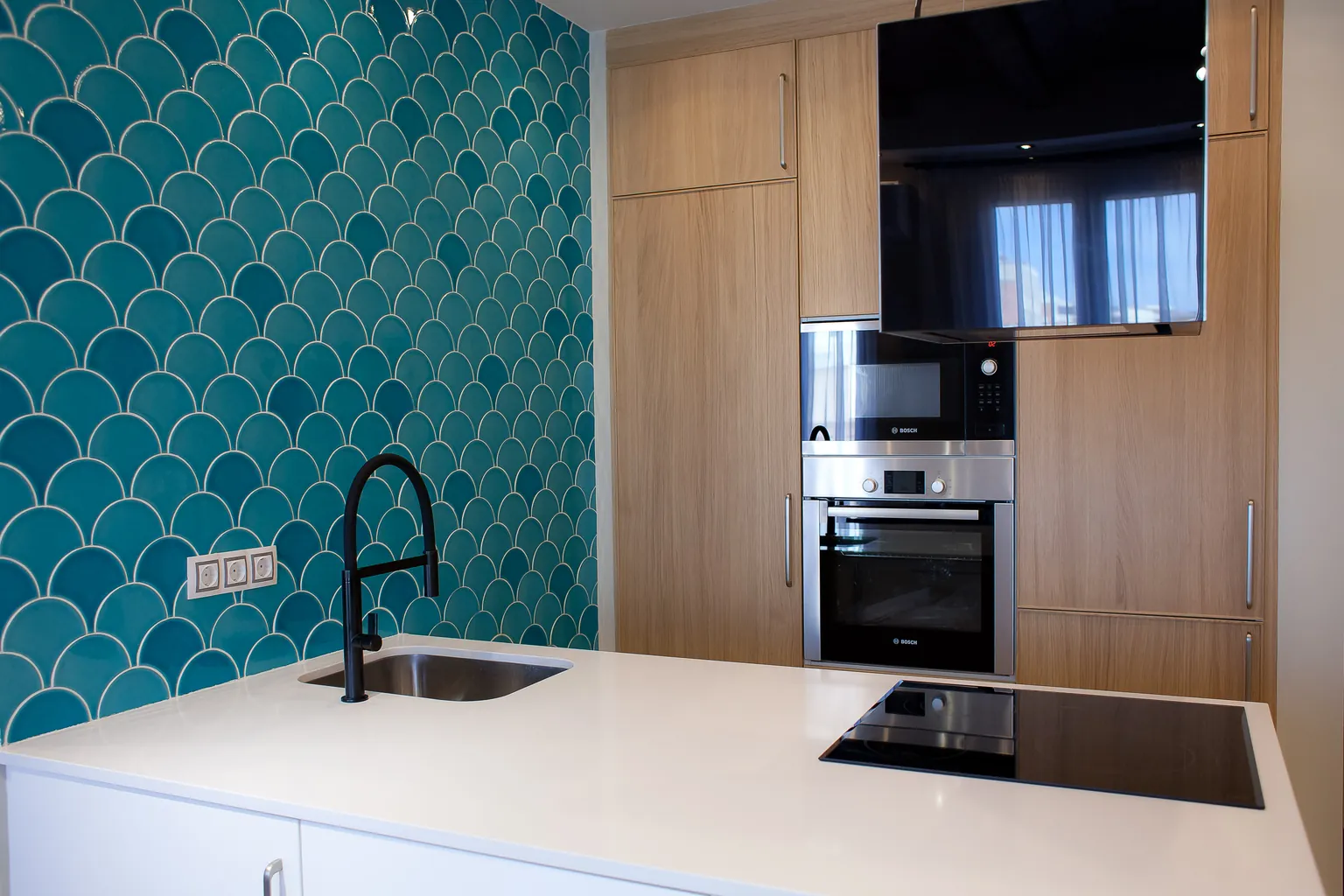 Cocina con azulejos turquesa en forma de escama de pez, isla blanca con fregadero y grifo negro, placa de inducción y columna de aspecto madera con horno y microondas integrados