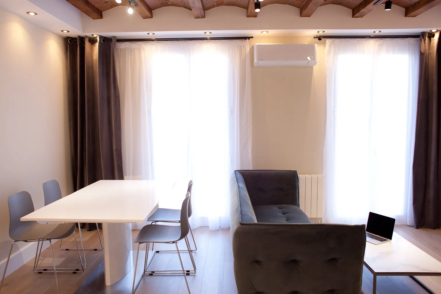 Comedor con mesa blanca y sillas grises junto al sofá, cortinas claras y techo de vigas de madera