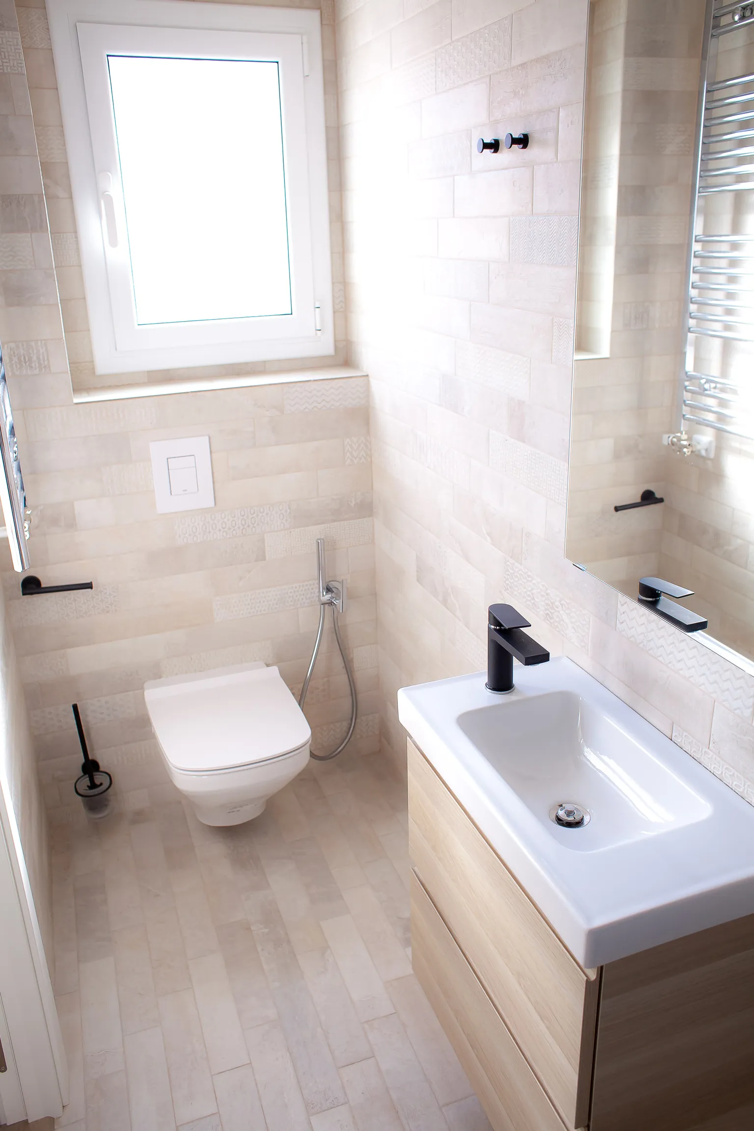 Segundo baño con azulejos claros de efecto piedra, inodoro suspendido, lavabo sobre mueble de madera, grifería negra y toallero