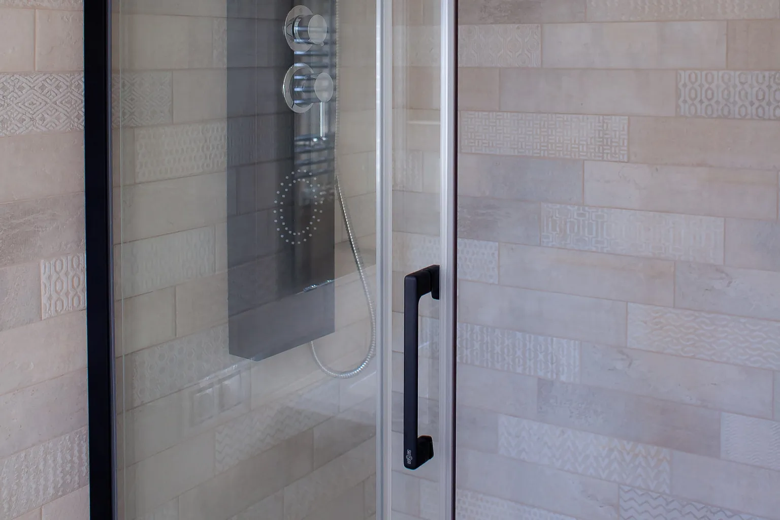 Detalle de la mampara de ducha con perfil negro, columna de hidromasaje y azulejos beige con relieves decorativos
