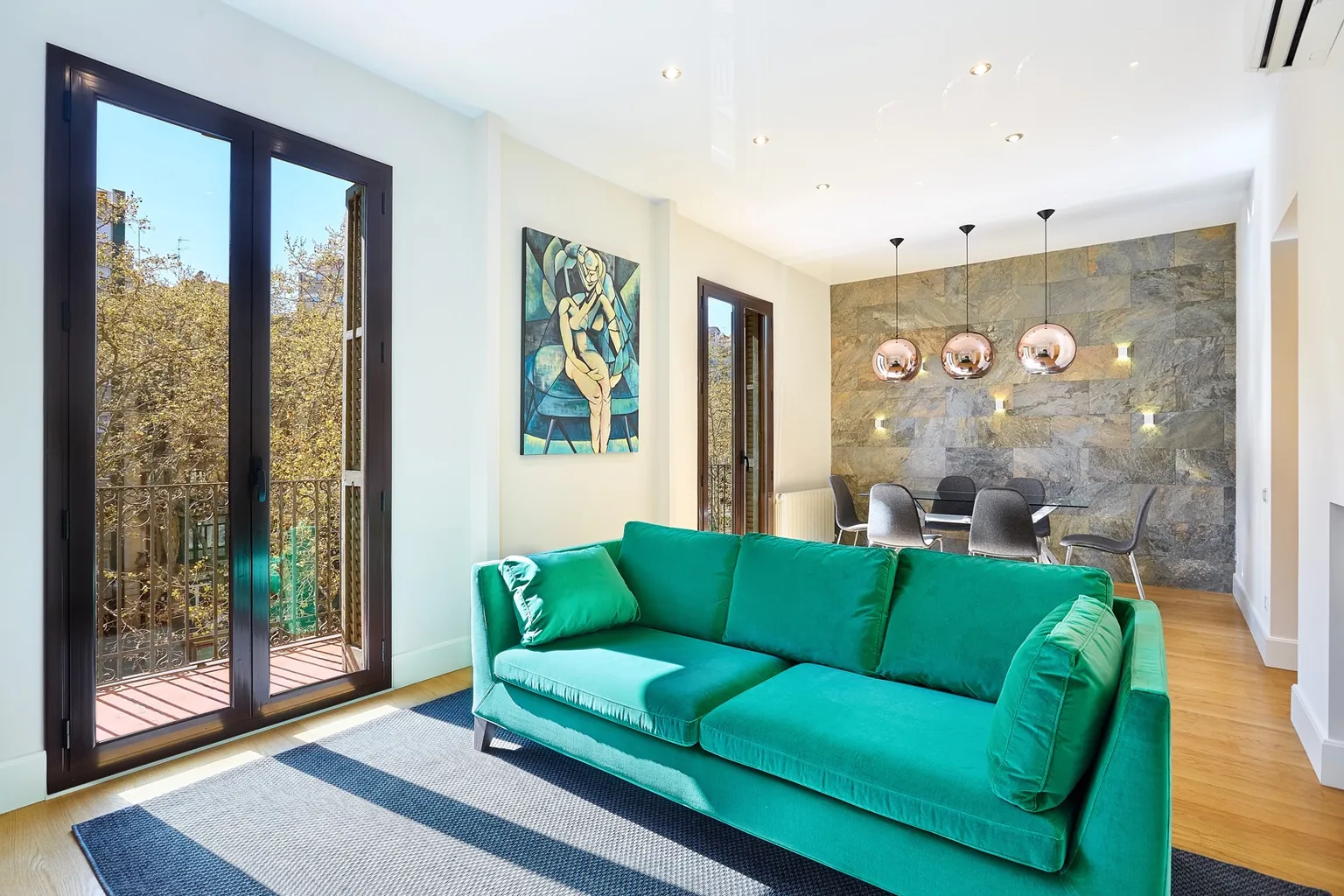 Living room of a renovated apartment in Barcelona with a green velvet sofa, wrought-iron balconies, copper pendant lamps and a stone-look wall in the dining area