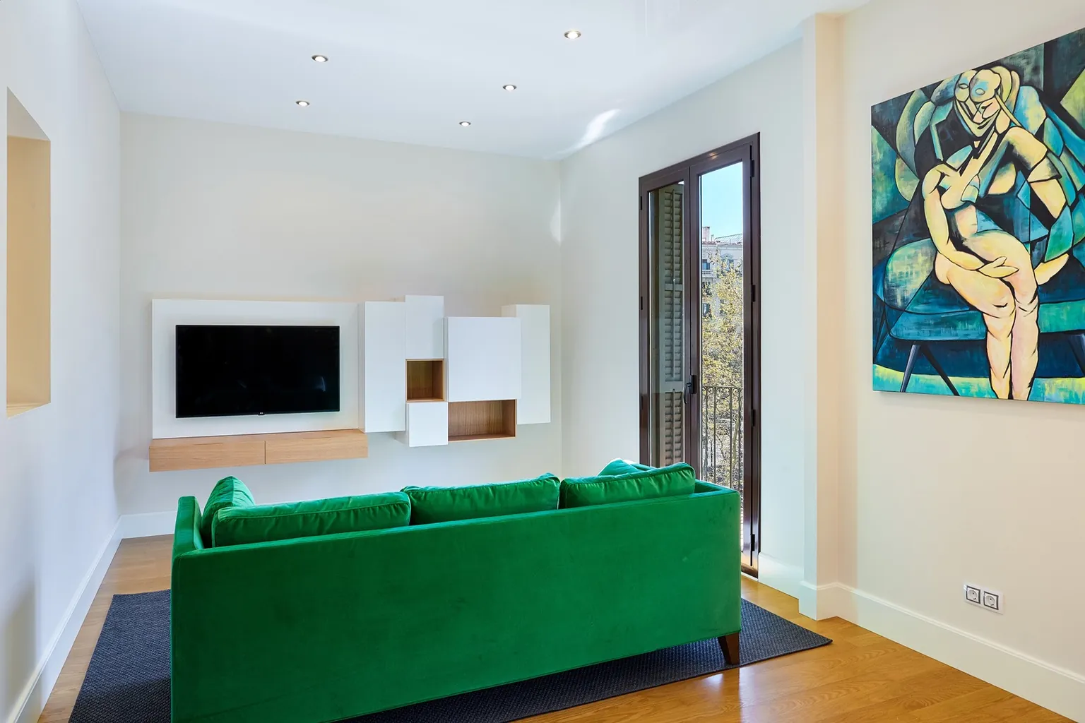 Salón con sofá verde, mueble de televisión blanco de módulos, balcón con contraventanas y cuadro de gran formato