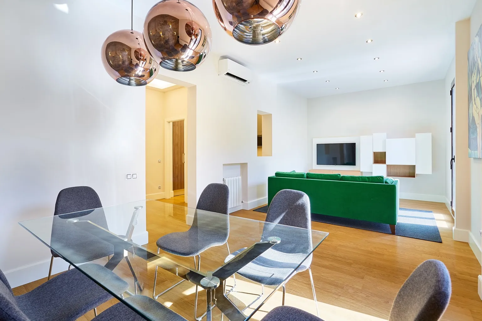 Comedor con mesa de vidrio, sillas grises y tres lámparas colgantes de cobre, con el salón y el sofá verde al fondo