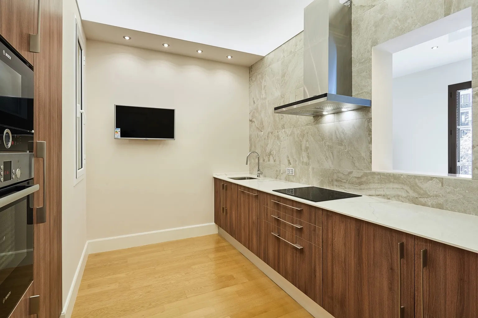 Cocina independiente con muebles de aspecto nogal, frontal y encimera de efecto mármol, campana de acero y televisor en la pared