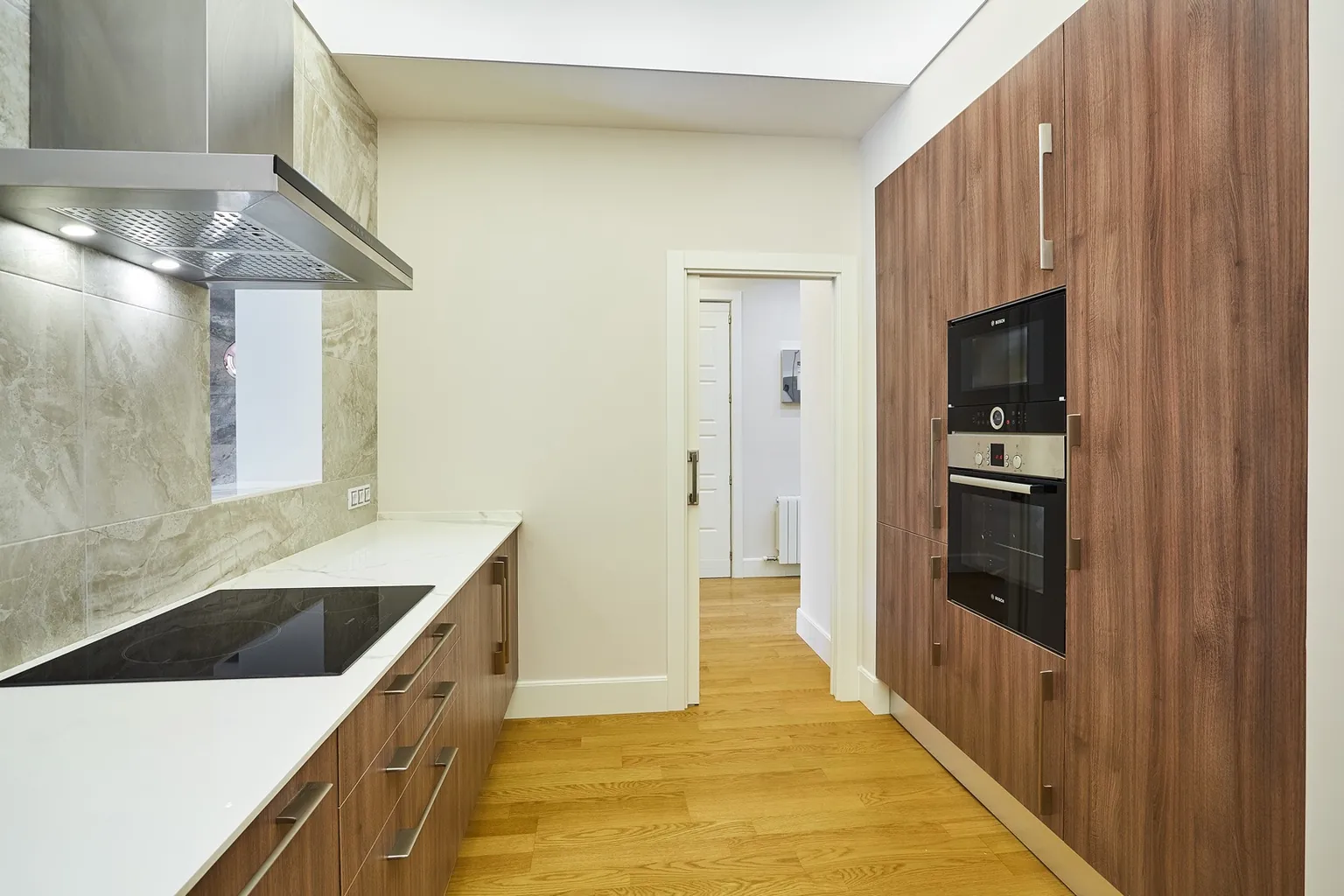 Cocina con placa de inducción, columna de nogal con horno y microondas integrados y puerta hacia el pasillo con parquet de roble