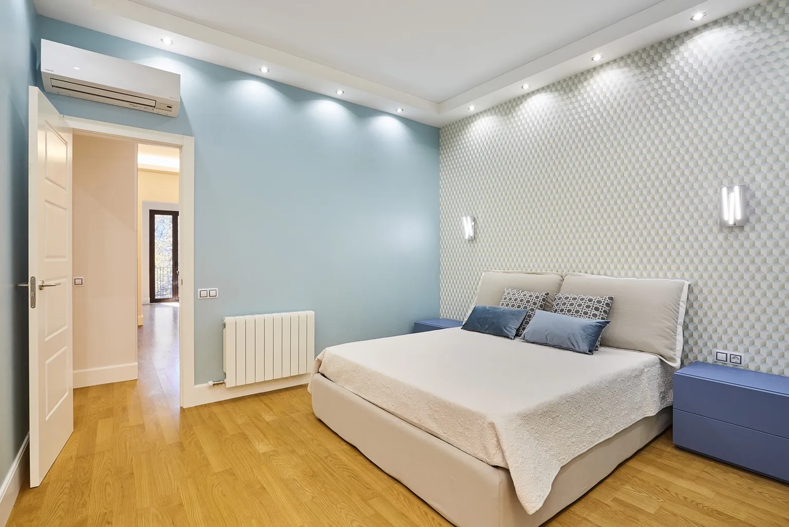 Dormitorio principal con pared azul, papel pintado geométrico tras el cabecero, apliques lineales, mesitas azules y parquet de roble