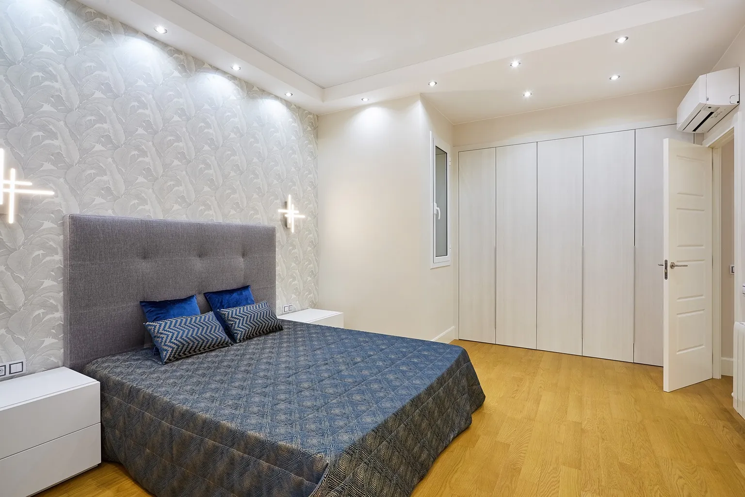 Segundo dormitorio con papel pintado de hojas, cabecero tapizado gris, apliques de luz lineal y armario blanco de puertas plegables