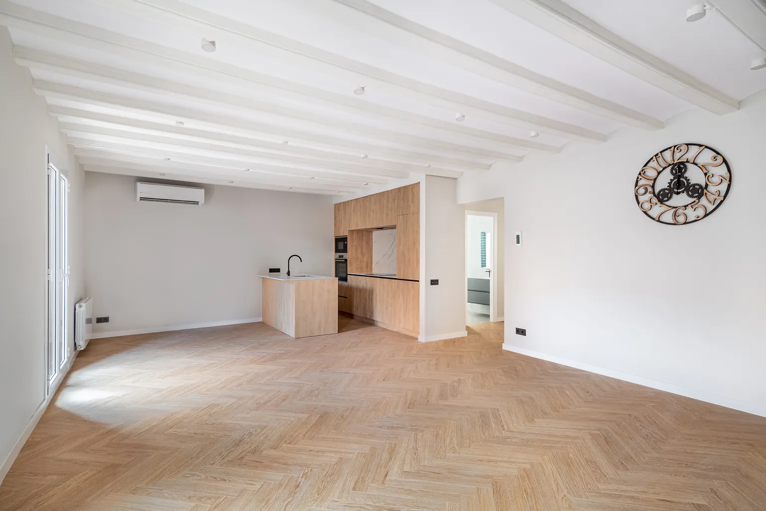 Living room with open kitchen in a renovated Gothic Quarter apartment in Barcelona: restored beamed ceilings painted white, herringbone flooring and wood-look units