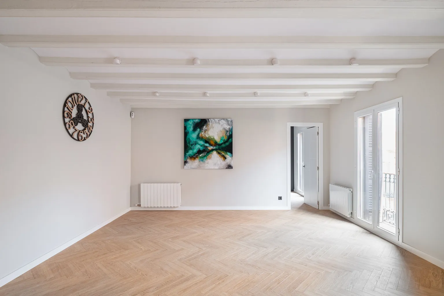 Living room with an abstract painting, two white radiators, beamed ceiling and a door onto the balcony with views over the neighbourhood