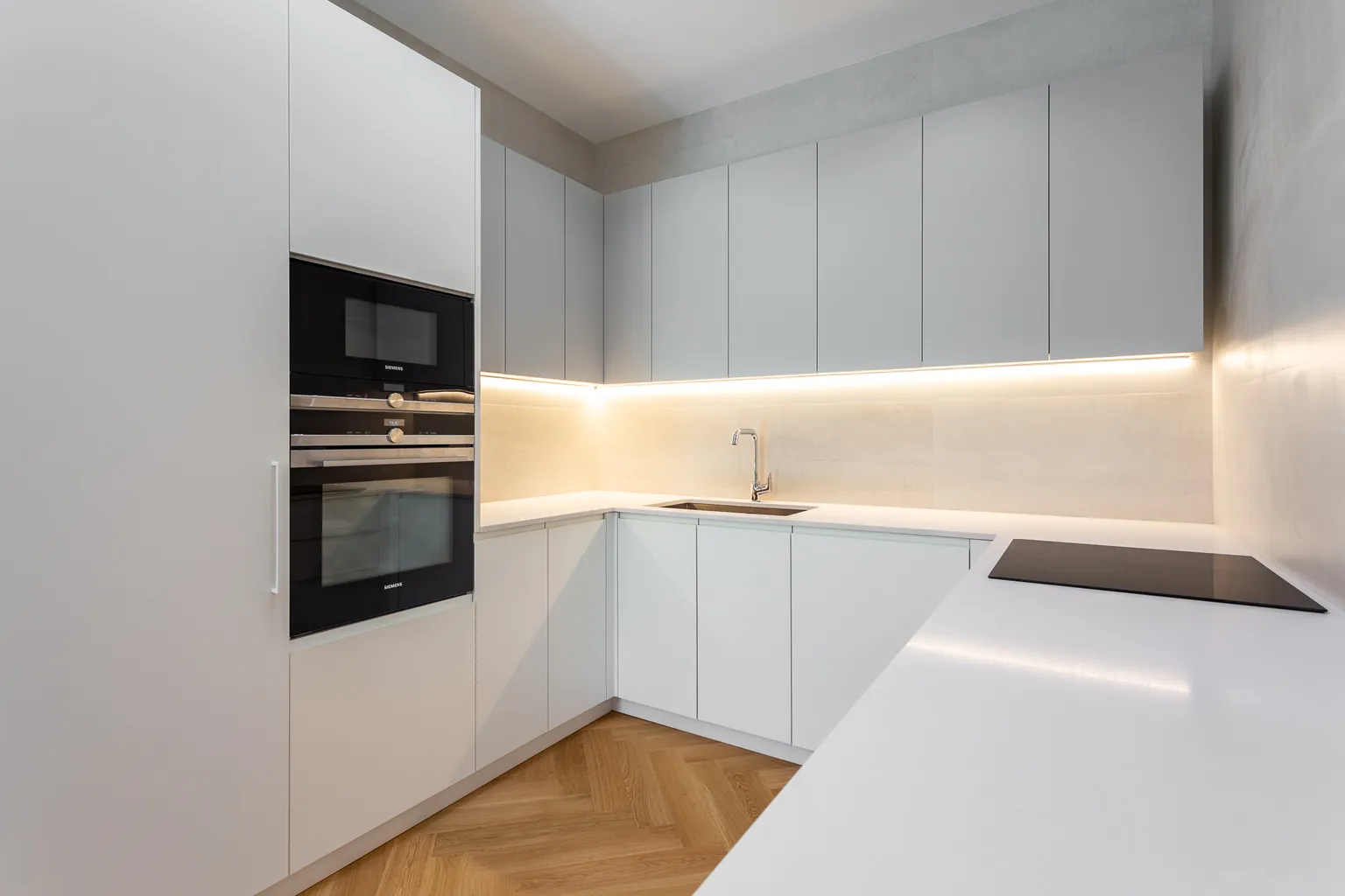 Cuisine blanche sans poignées avec colonne de fours encastrés, plan de travail blanc et éclairage LED sous les meubles hauts