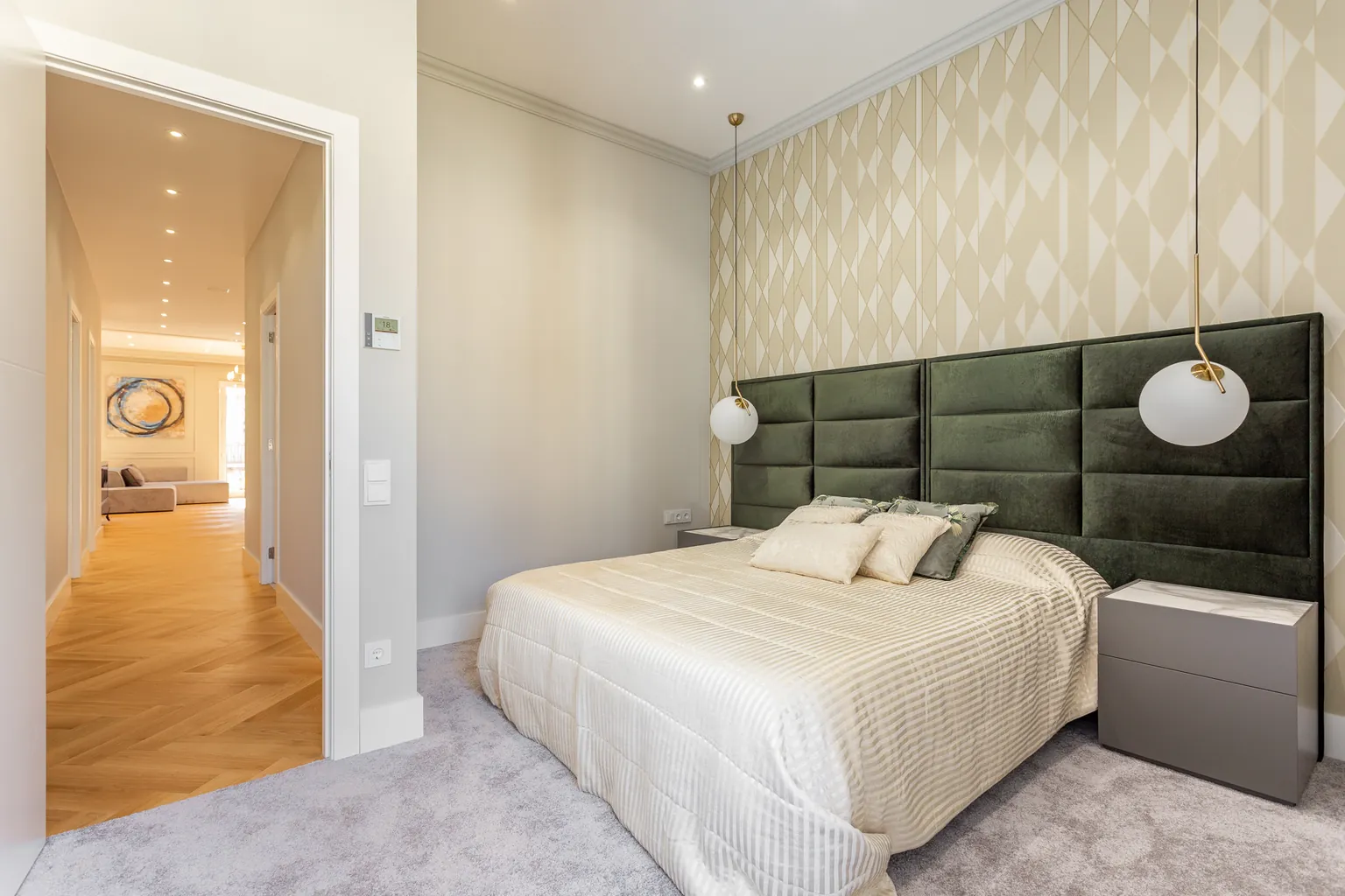 Chambre principale avec tête de lit capitonnée verte, papier peint géométrique et vue sur le couloir au parquet à bâtons rompus