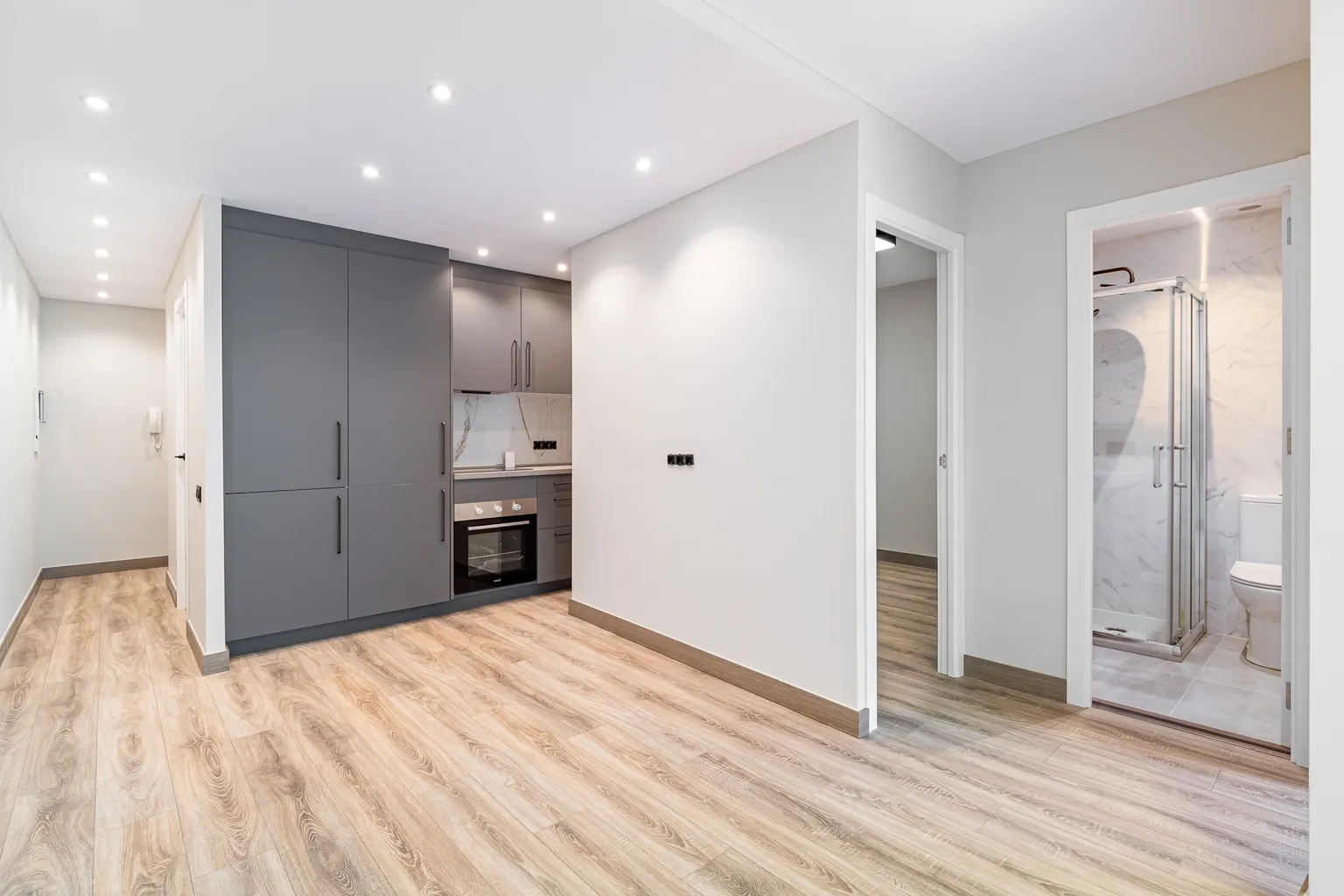 Living room of a renovated apartment in Gavà with an open grey kitchen, wood-look flooring, recessed spotlights and open doors to the bedroom and the bathroom