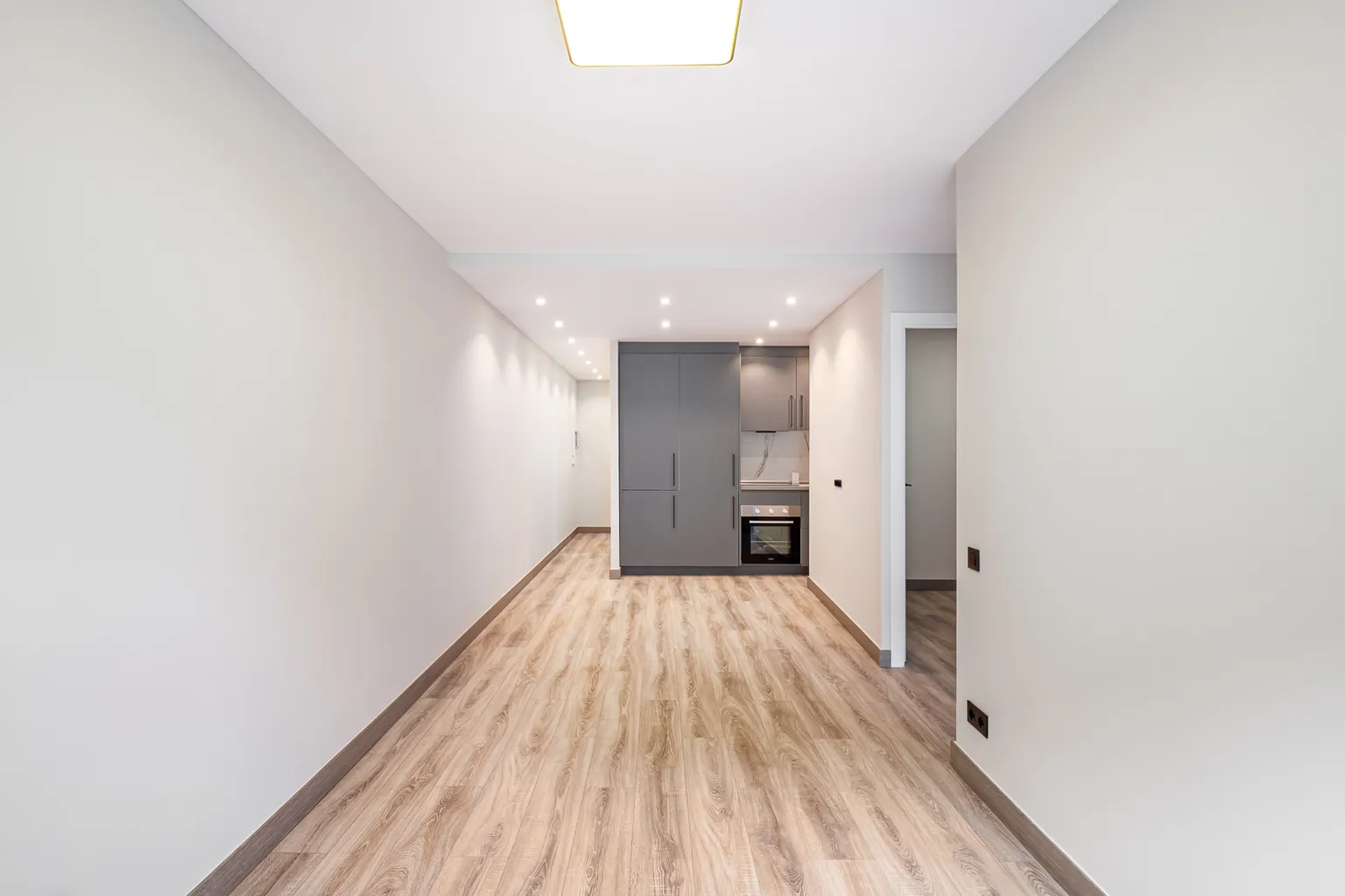 Salón alargado con plafón de techo, paredes blancas, suelo laminado de aspecto madera y la cocina gris al fondo