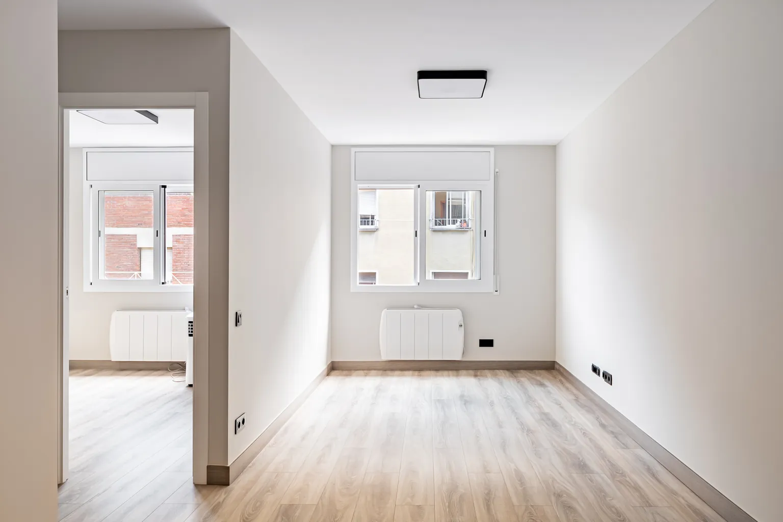 Salón con ventana, radiador eléctrico, plafón negro y paso al dormitorio con otra ventana