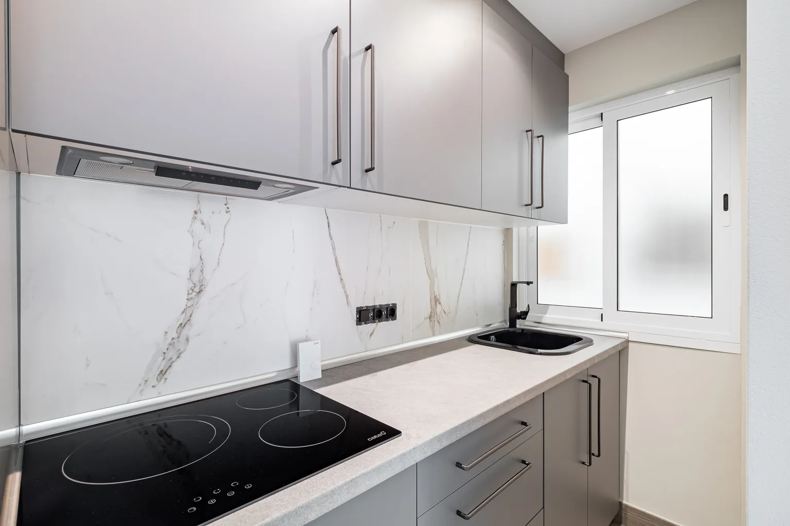 Detalle de la cocina gris con frontal de efecto mármol, placa de inducción, fregadero y grifo negros y ventana de vidrio translúcido