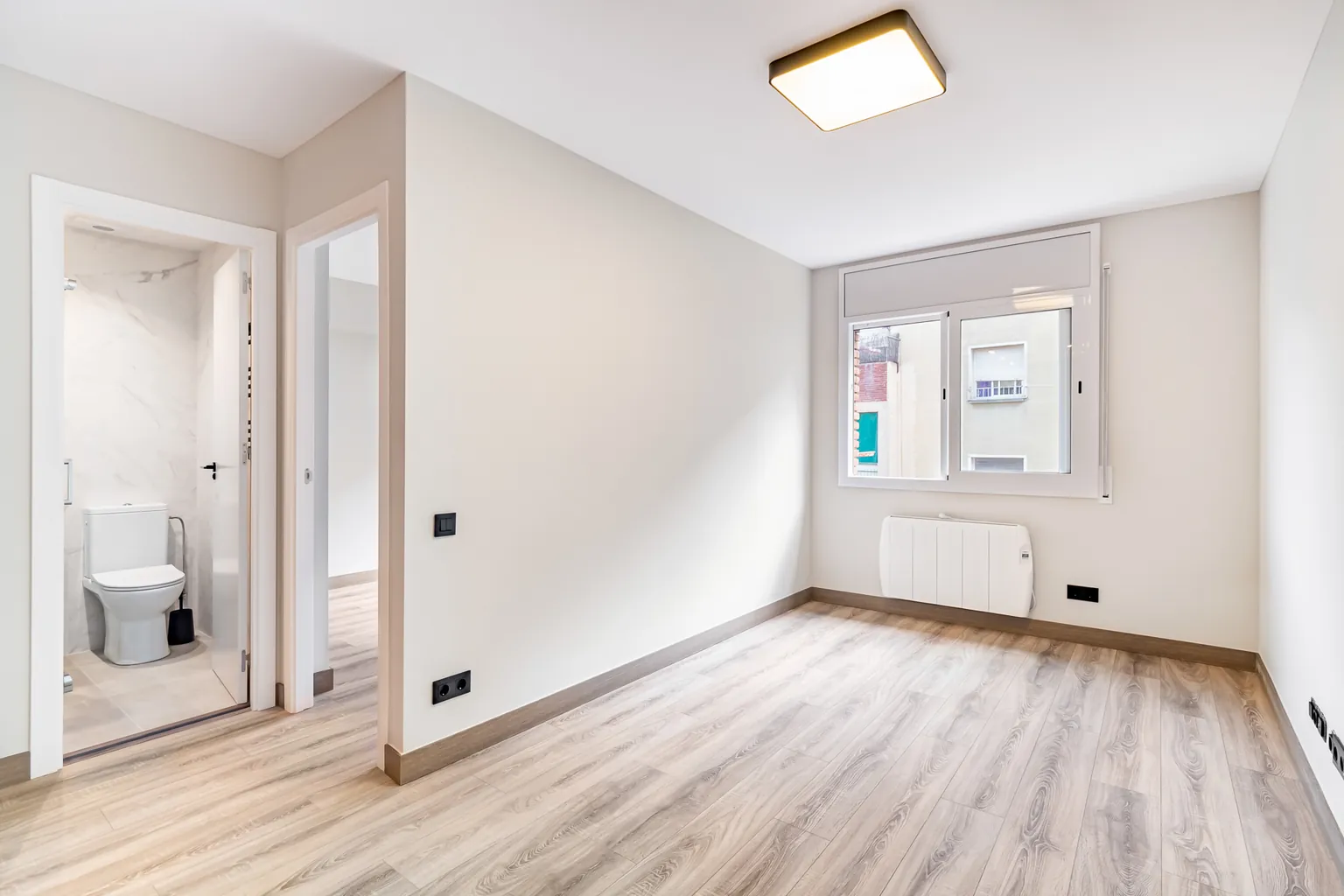 Dormitorio con ventana, plafón de techo, radiador eléctrico y puertas abiertas al baño y al pasillo