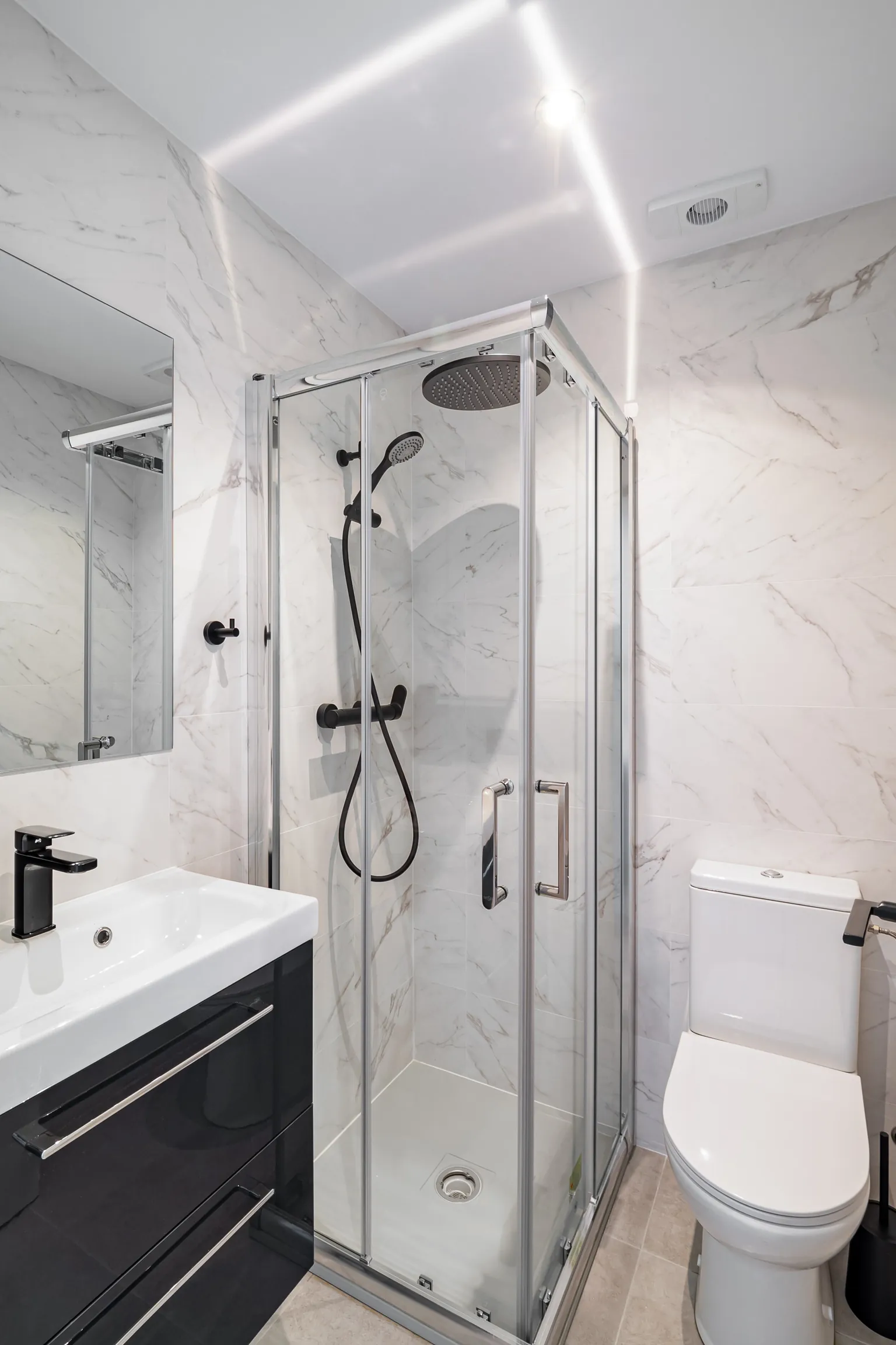 Baño con porcelánico efecto mármol blanco, ducha de esquina con mampara, grifería negra, mueble negro e inodoro