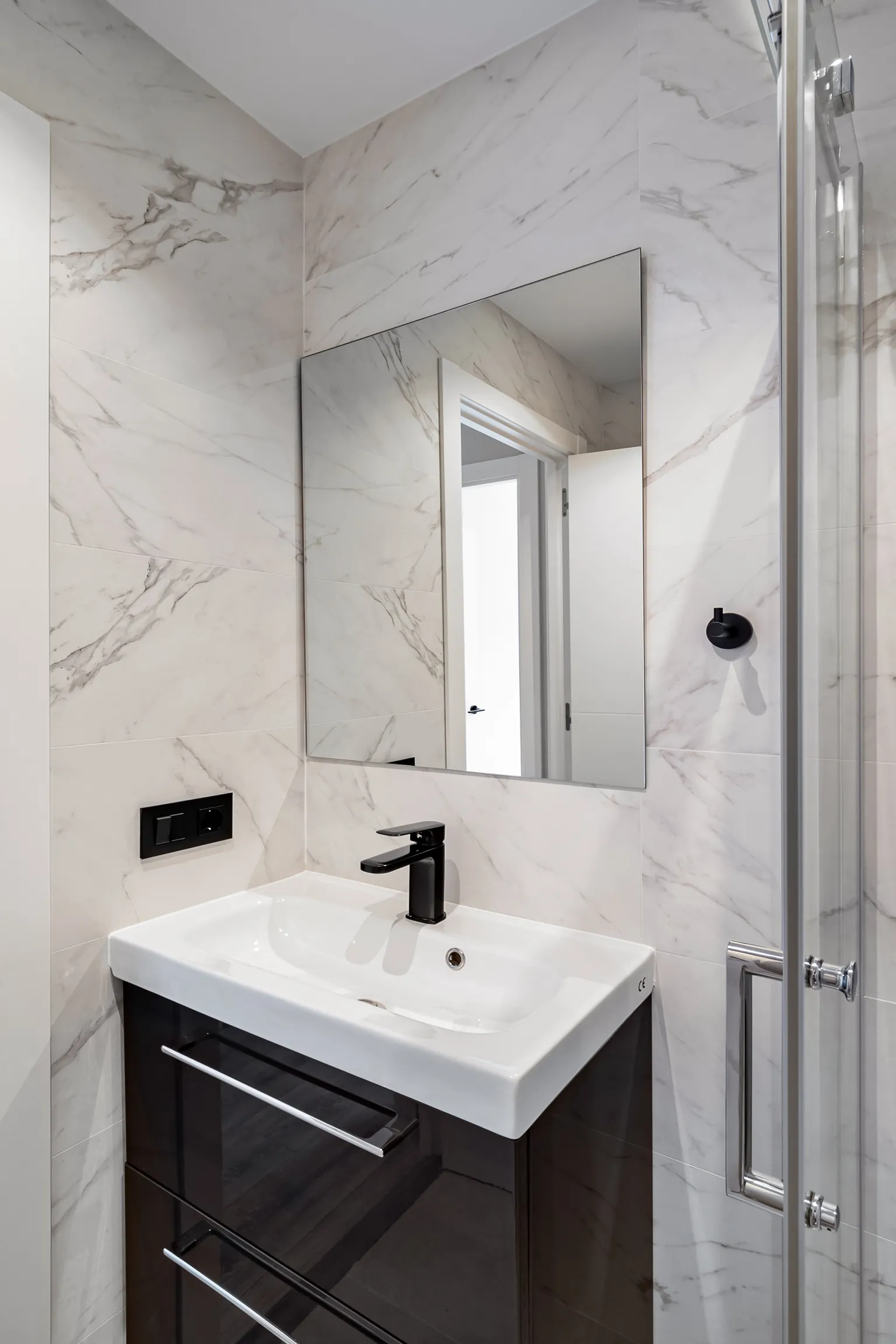 Lavabo sobre mueble negro brillante con grifo negro y espejo en un baño de porcelánico efecto mármol