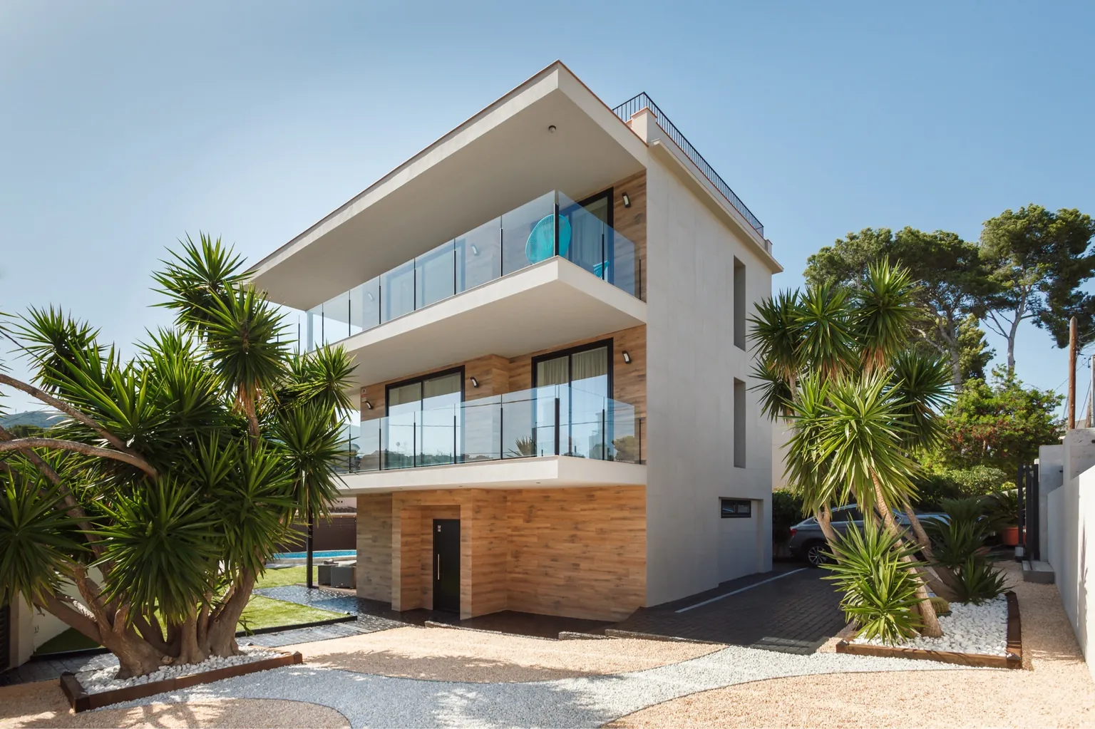 Wyremontowany dom w Castelldefels: nowoczesna trzykondygnacyjna elewacja z tarasami ze szklanymi balustradami, okładziną o wyglądzie drewna, palmami i żwirowym ogrodem
